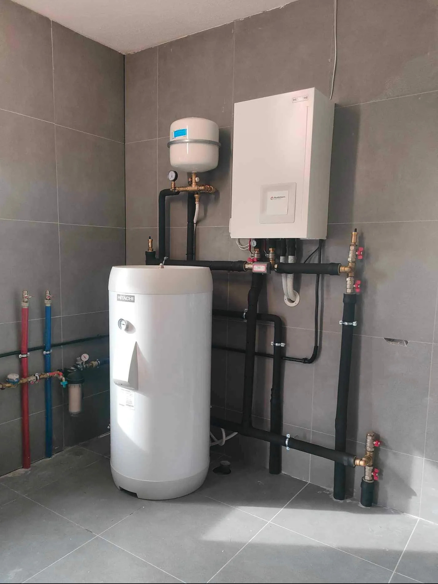 Biały zasobnik ciepłej wody Hitachi i kocioł Buderus na tle szarych płytek. Czarny rurociąg z zaworami. Kompaktowa instalacja grzewcza w pomieszczeniu technicznym.