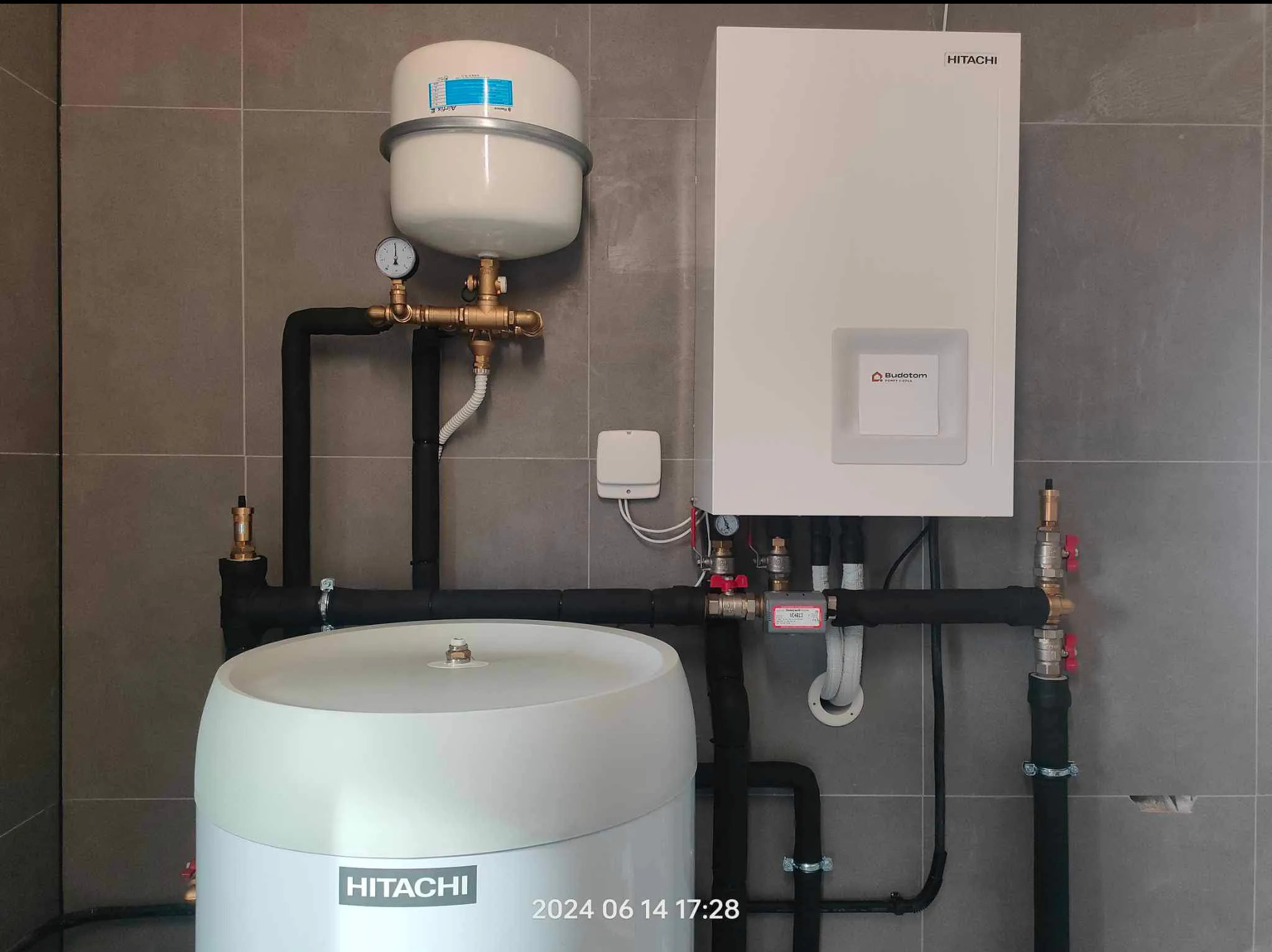 Zestaw urządzeń Hitachi: pompa ciepła z zbiornikiem buforowym i naczyniem wzbiorczym, instalacja w szarym pomieszczeniu, widoczne rury i zawory.