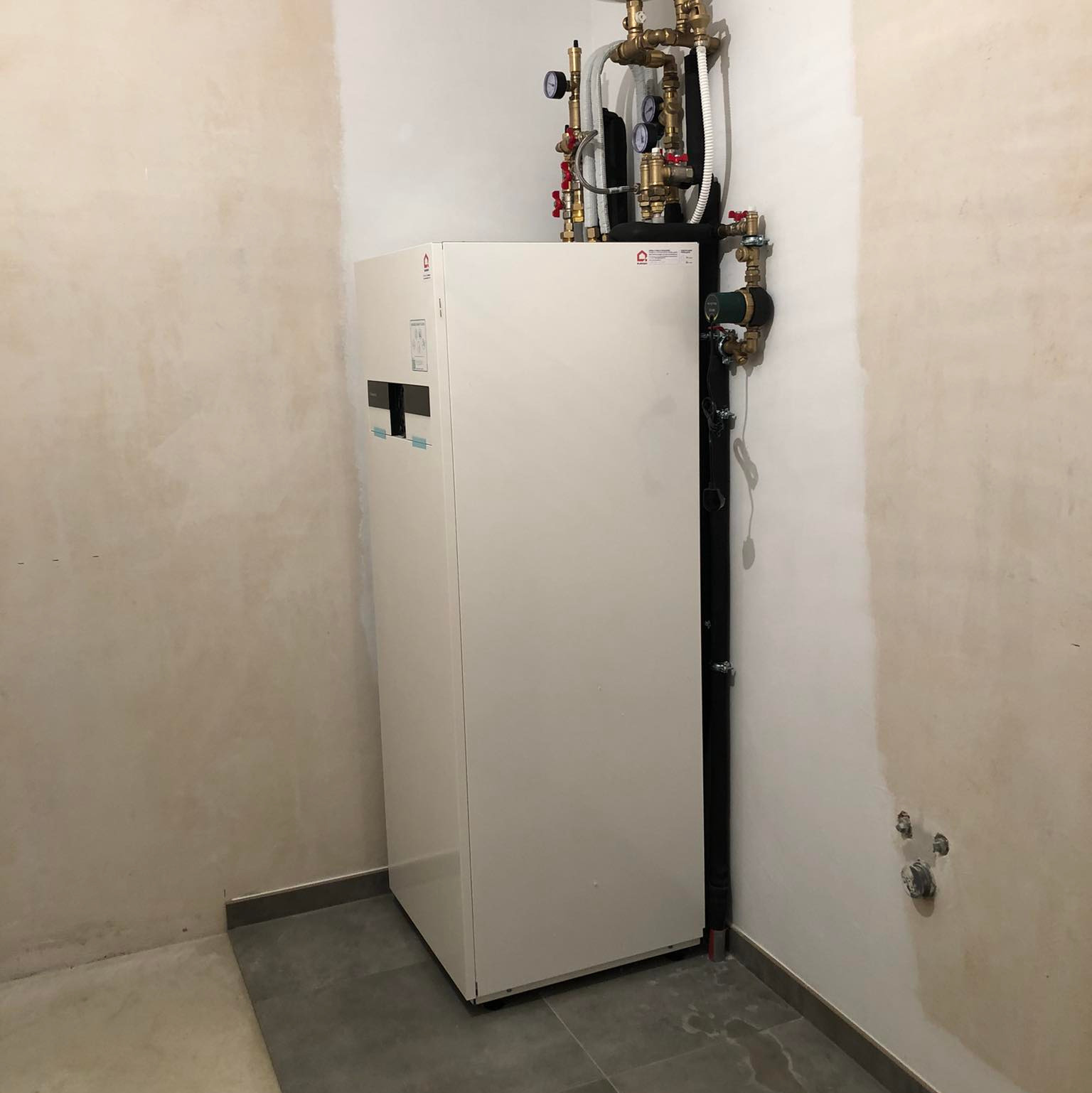 Biała pompa ciepła z podłączonymi rurami i zaworami w surowym pomieszczeniu z szarymi płytkami podłogowymi. Instalacja w trakcie realizacji.