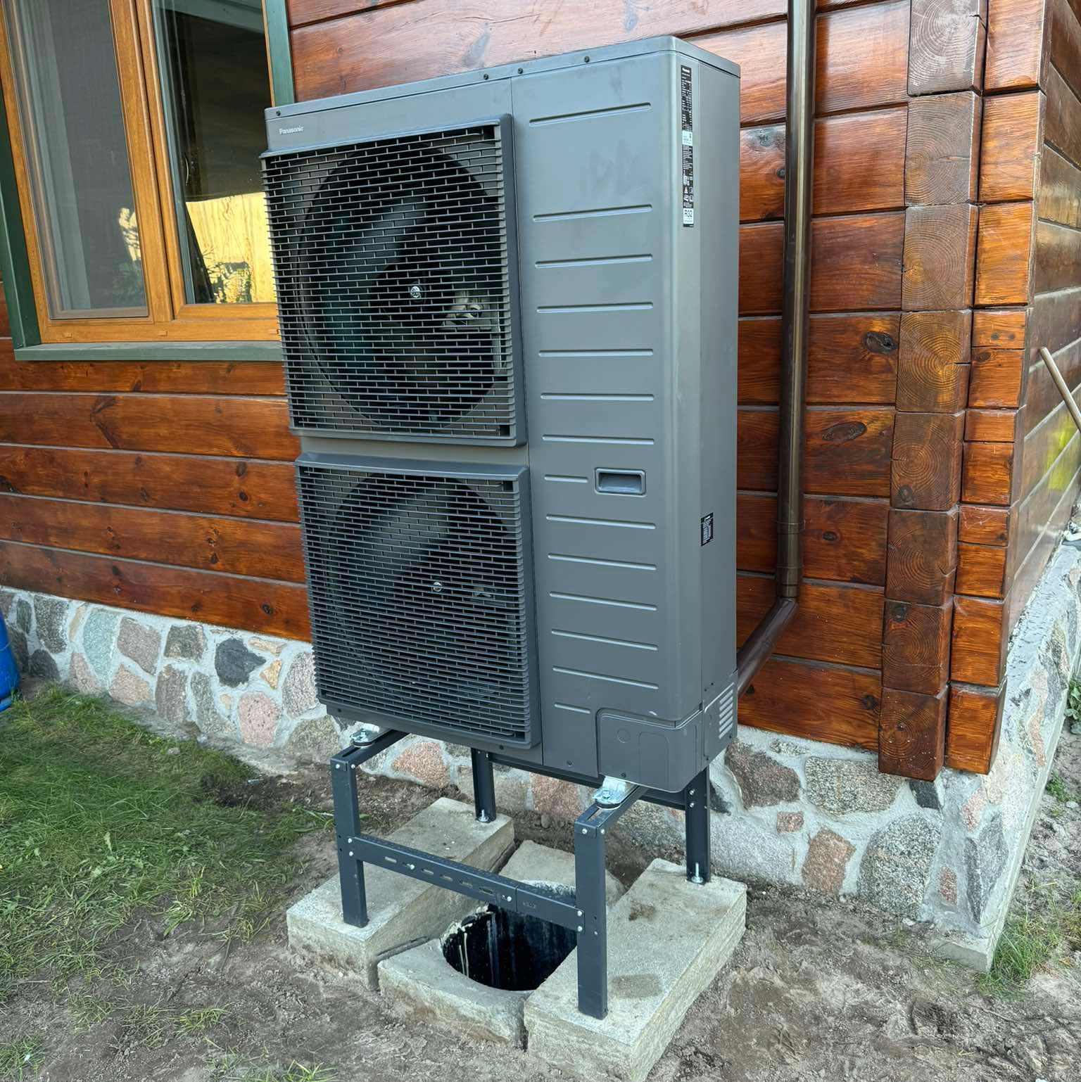 Szara pompa ciepła Panasonic na metalowym stelażu, zamontowana przy drewnianym domu z kamienną podmurówką. Widoczna rura odprowadzająca skropliny.