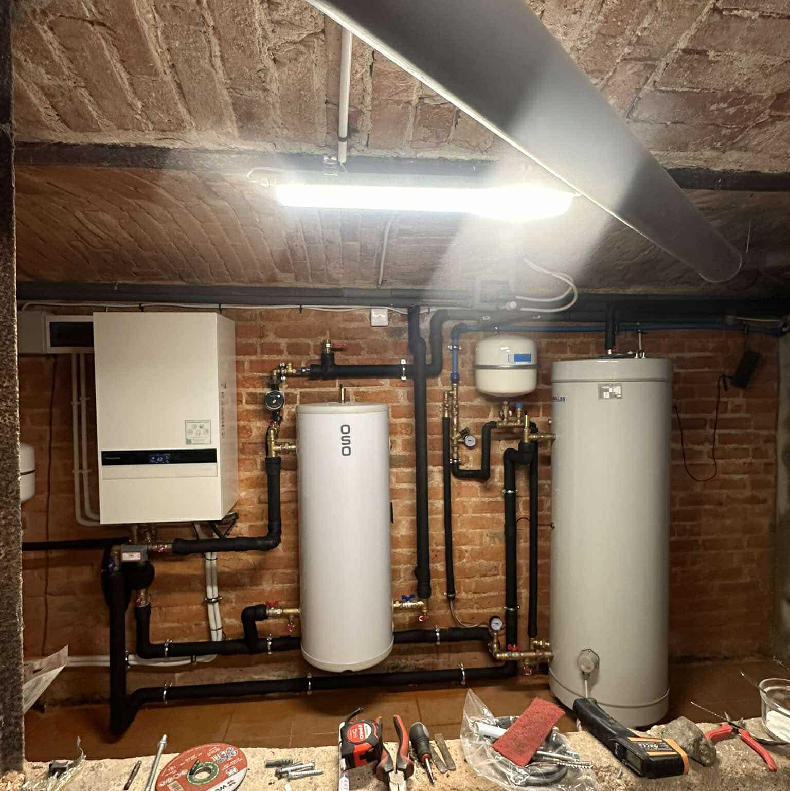Instalacja grzewcza w piwnicy z ceglanym sklepieniem: biały piec, dwa cylindryczne zbiorniki OSO, czarne rury. Na podłodze narzędzia monterskie.