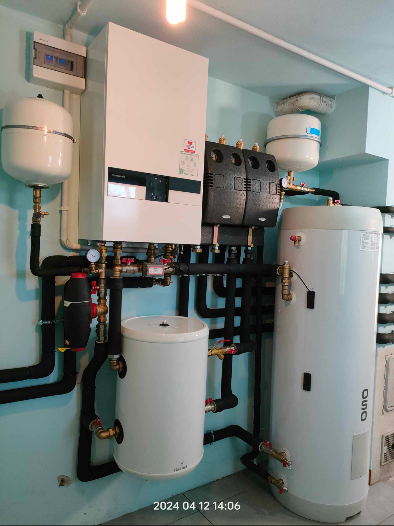 Złożona instalacja grzewcza z kotłem, zasobnikami i rurami, zamontowana na ścianie o jasnoniebieskim odcieniu, z widocznymi elementami sterującymi i zabezpieczającymi.