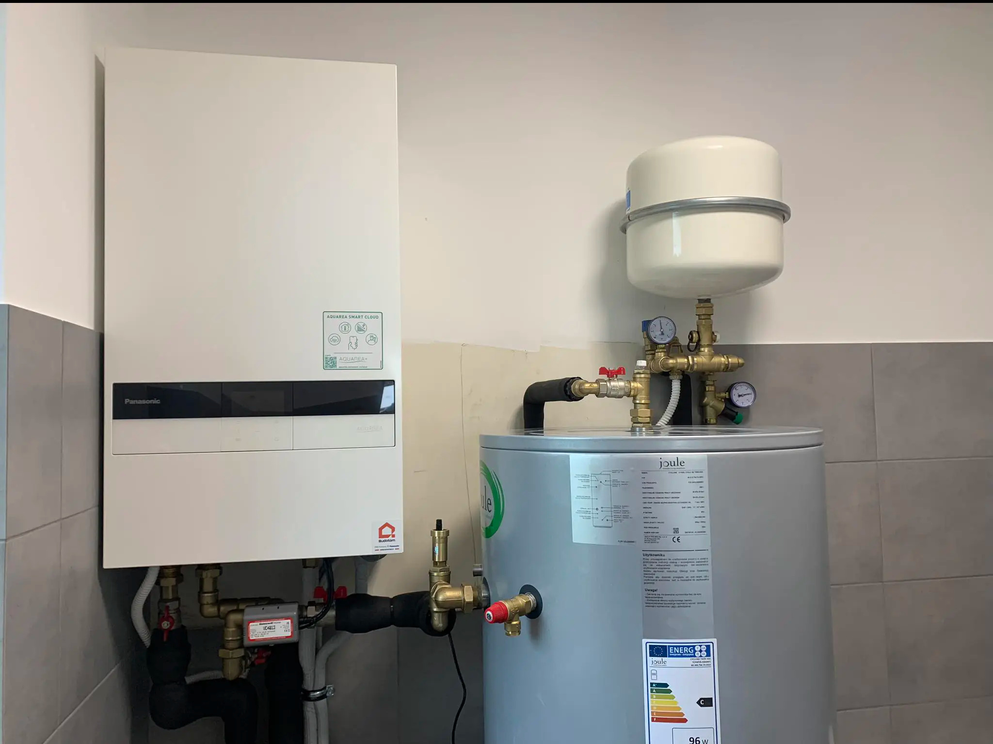 Biały piec Panasonic Aquarea Smart Cloud i srebrny bojler Joule z czarnymi rurami, manometrami i zaworami w pomieszczeniu z szarą glazurą.