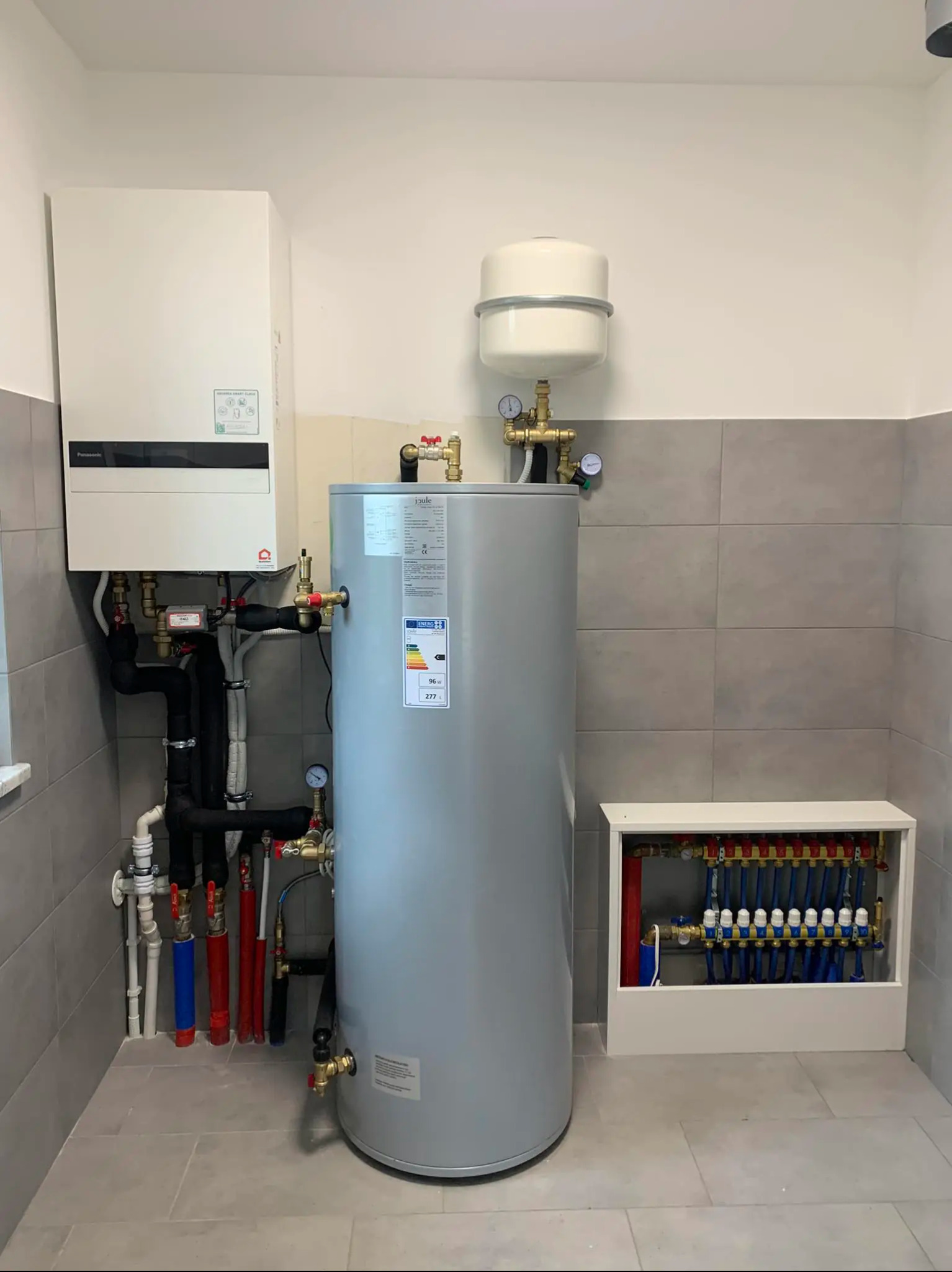 Biały piec grzewczy, szary zbiornik i rozdzielacz ogrzewania podłogowego w pomieszczeniu z szarą glazurą. Elementy instalacji grzewczej w kotłowni.