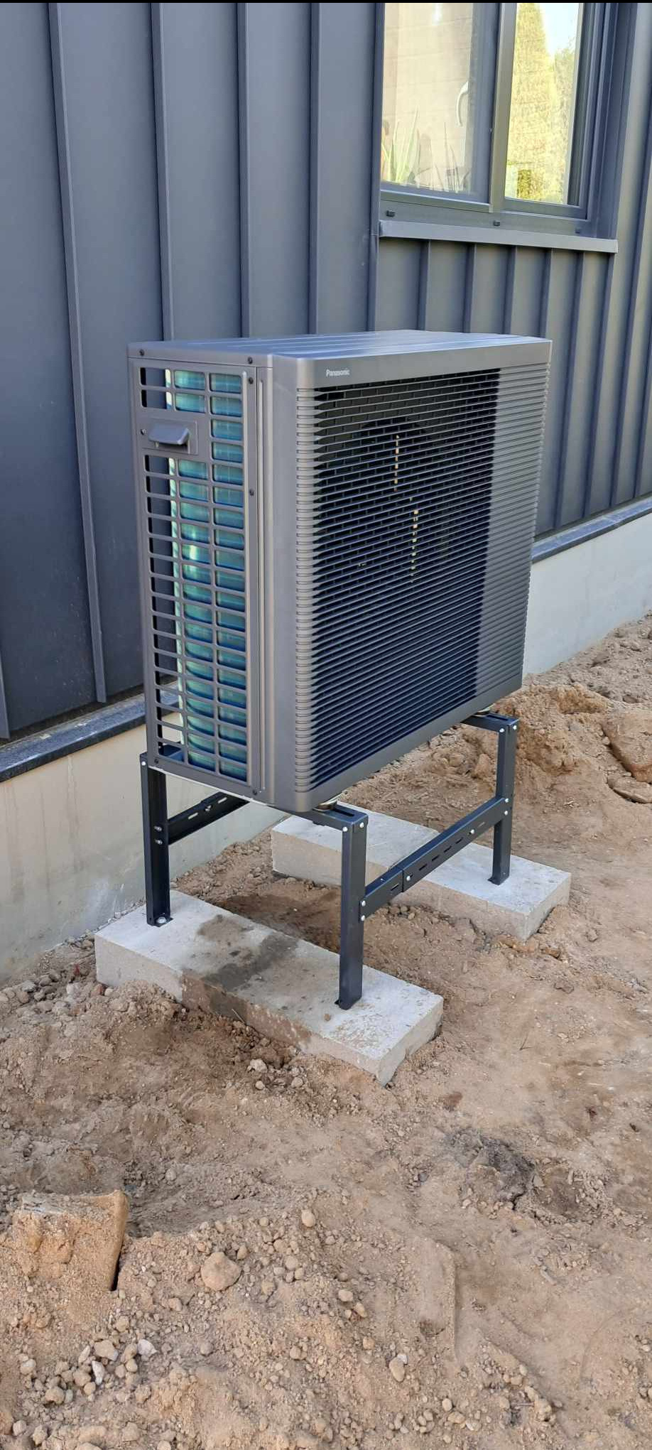 Szara pompa ciepła Panasonic zamontowana na czarnym stelażu, umieszczonym na betonowych płytach. Tło stanowi szara elewacja budynku z oknem.