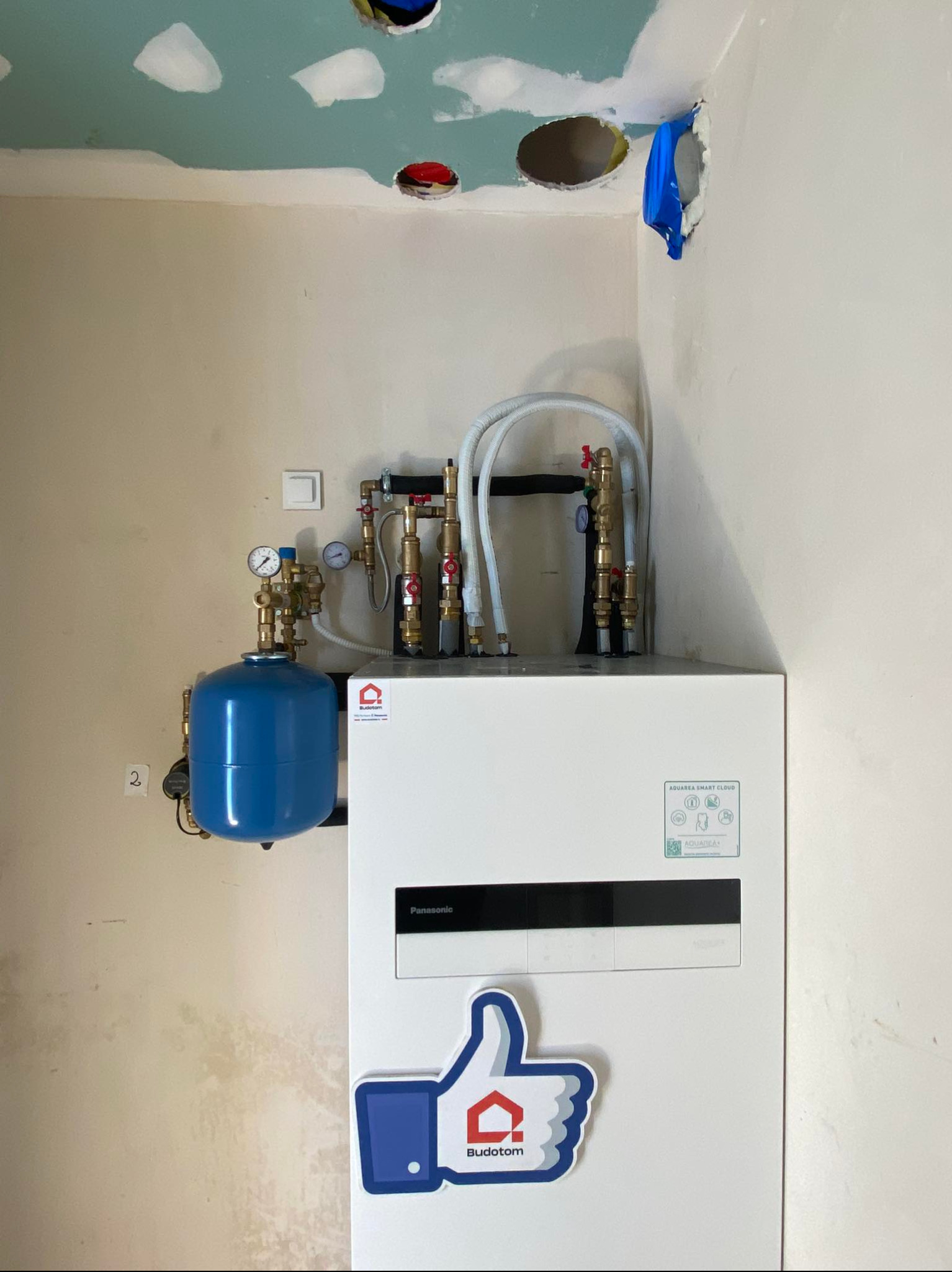 Biała pompa ciepła Panasonic z niebieskim zbiornikiem wyrównawczym i rurami miedzianymi w rogu pokoju w trakcie wykańczania. Naklejka z logo firmy.