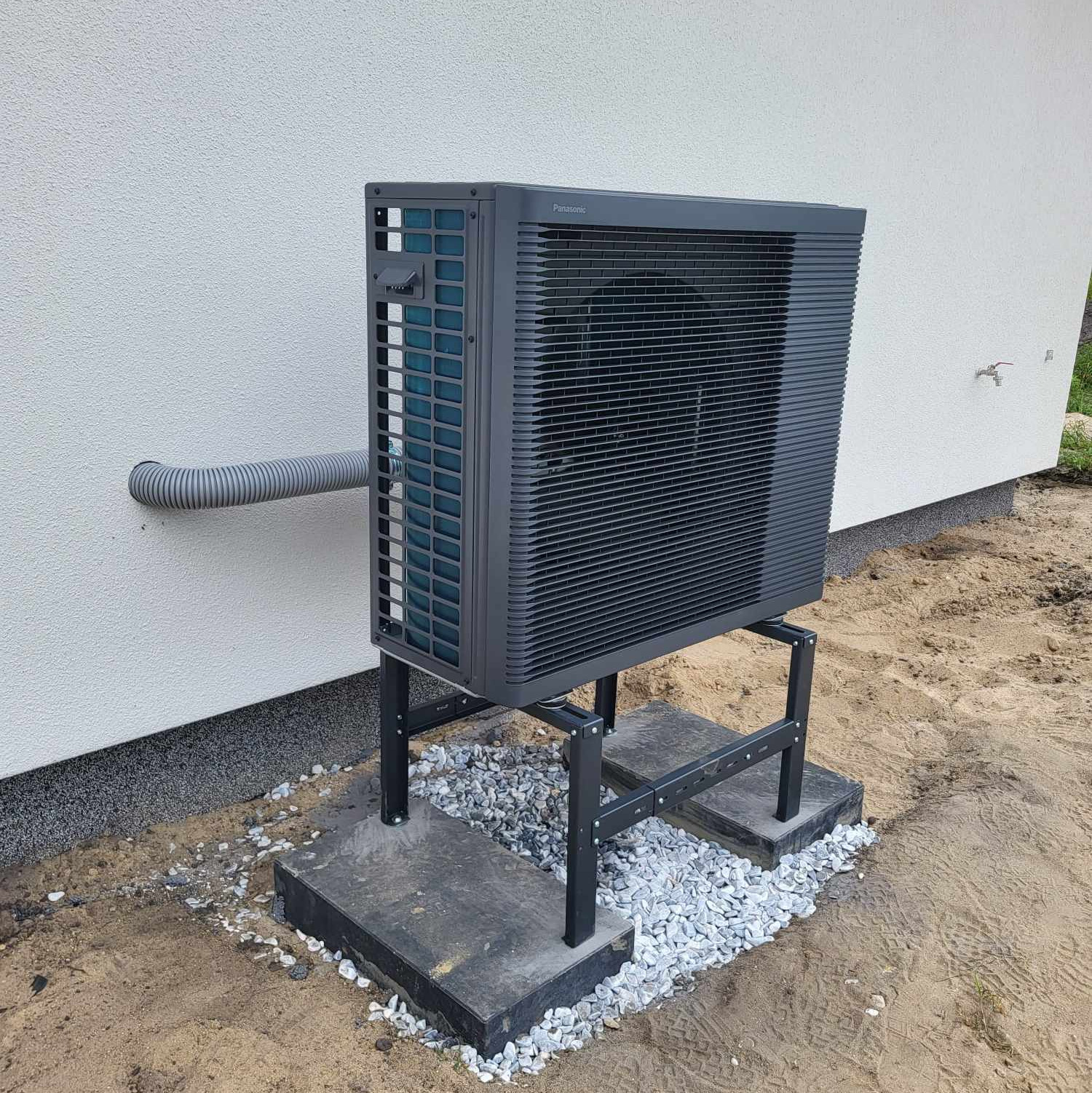 Nowoczesna, grafitowa pompa ciepła Panasonic Aquarea zamontowana na betonowej podstawie z kamieniami, przy jasnej ścianie budynku z rurą przyłączeniową.