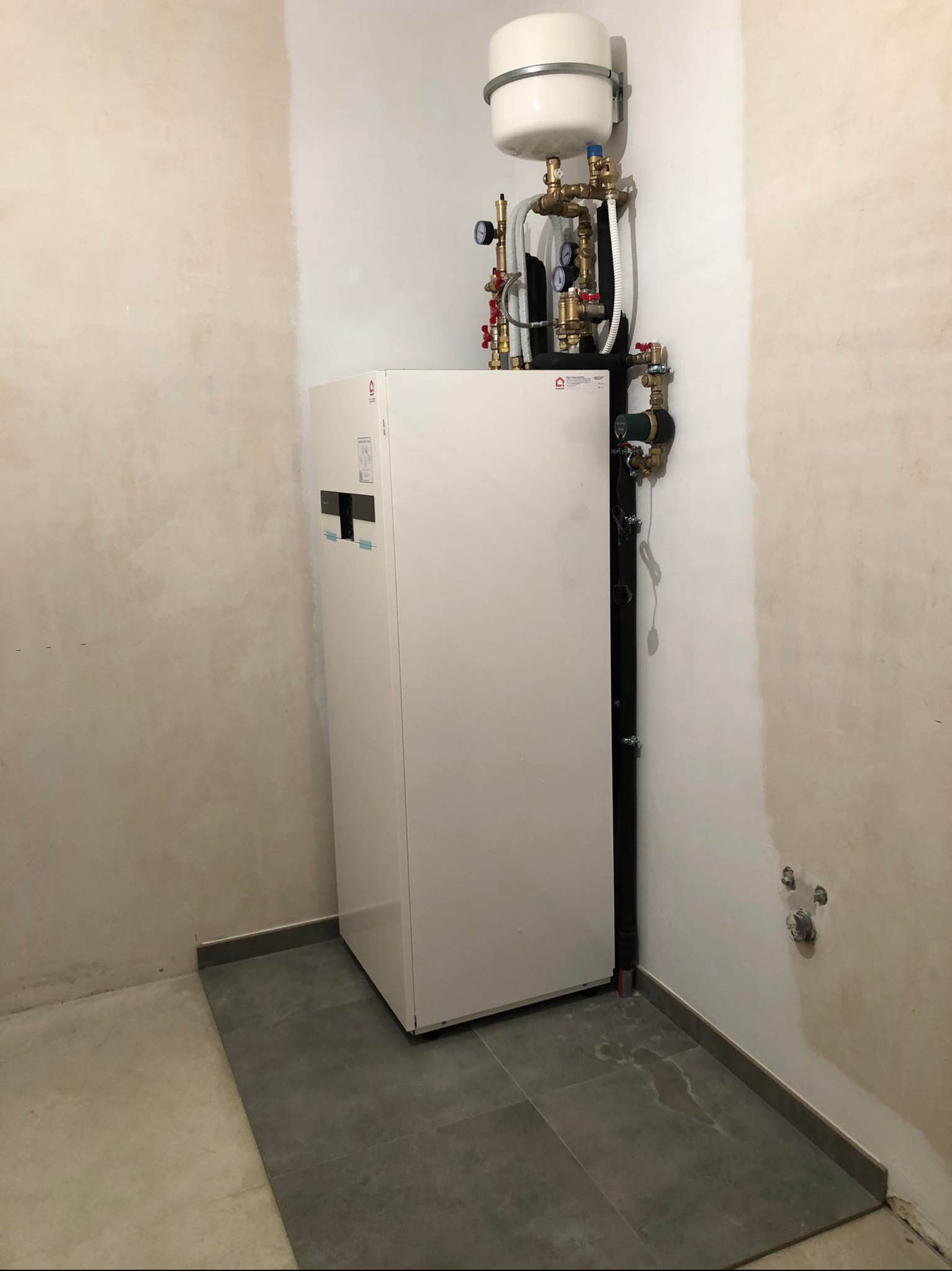 Biała pompa ciepła w rogu pomieszczenia z szarą podłogą i surowymi ścianami. Widoczne rury i zawory. Realizacja instalacji w trakcie wykończenia.