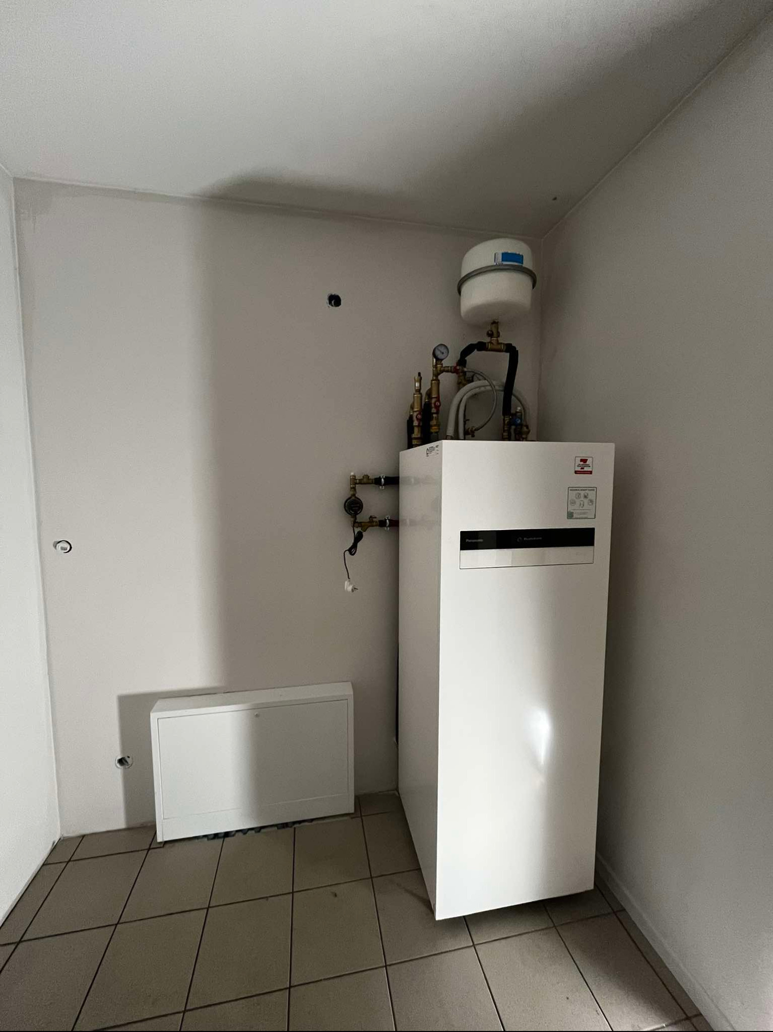 Biała pompa ciepła Panasonic Aquarea w rogu pomieszczenia z płytkami. Widoczne rury, manometry i zbiornik wyrównawczy. Obok biała szafka instalacyjna.