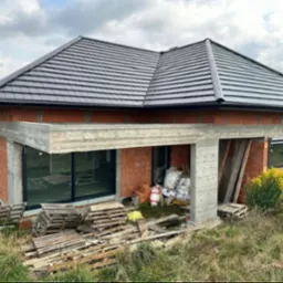 Dom w trakcie budowy: czerwona cegła, betonowe nadproża, grafitowy dach. Widoczne palety i materiały budowlane. Krajobraz w tle.