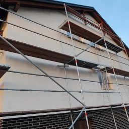 Rafał Kędziora - Dom jednorodzinny w trakcie malowania elewacji, widoczne rusztowania i świeżo pomalowana ściana w jasnym odcieniu. Dolna część elewacji wykończona ciemną cegłą.