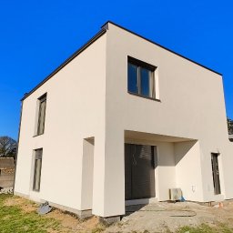 Rafał Kędziora - Nowoczesny, dwukondygnacyjny dom z jasną elewacją i ciemnymi oknami, na tle błękitnego nieba i zielonego trawnika. W tle widoczny traktor i betonowe ogrodzenie.