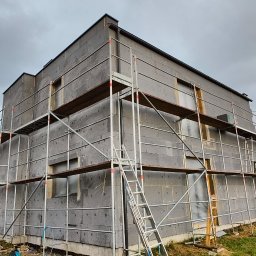 Rafał Kędziora - Dom w trakcie ocieplania, pokryty szarym styropianem, otoczony rusztowaniami. Widoczny fragment ogrodu i pochmurne niebo.