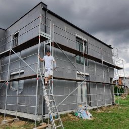 Rafał Kędziora - Dom w trakcie ocieplania, pokryty szarym styropianem, otoczony rusztowaniami. Pracownik na drabinie z tubą kleju. Pochmurne niebo w tle.