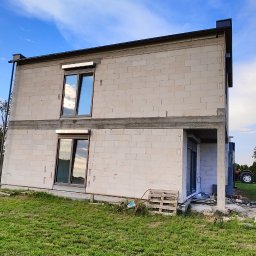 Rafał Kędziora - Dwukondygnacyjny dom w stanie surowym otwartym, z widocznymi oknami z roletami zewnętrznymi i niezakończoną elewacją z betonu komórkowego. Na trawniku paleta i narzędzia.