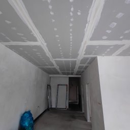 Rafał Kędziora - Sufit z płyt kartonowo-gipsowych, świeżo po szpachlowaniu, z widocznymi łączeniami i punktowymi śladami po szpachli. W tle widoczne ściany w trakcie prac wykończeniowych.