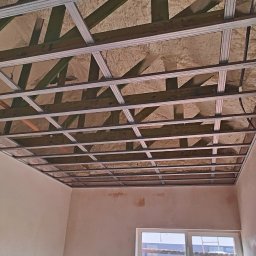 Rafał Kędziora - Sufit podwieszany z metalową konstrukcją i izolacją z pianki, widok z dołu w surowym wnętrzu. Ściany przygotowane do malowania, okno z pianką montażową.