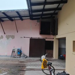 Rafał Kędziora - Budynek z fragmentami elewacji w trakcie malowania na beżowo, widoczne narzędzia malarskie i rower. Część budynku w kolorze różowym z odpadającym tynkiem.