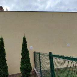 Rafał Kędziora - Jasna elewacja budynku z trzema tujami i zielonym ogrodzeniem. Widoczne detale: otwór w ścianie, kratki wentylacyjne. Pochmurne niebo w tle.