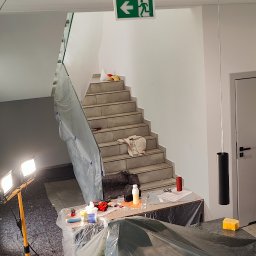 Rafał Kędziora - Przygotowania do malowania klatki schodowej: zabezpieczone schody folią, narzędzia na stole, oświetlenie robocze. Widoczny znak ewakuacyjny.