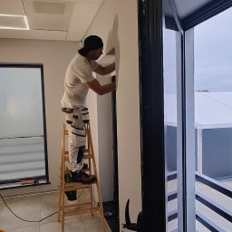 Rafał Kędziora - Pracownik na drabinie szpachluje ścianę w jasnym wnętrzu, widoczne okno balkonowe i sufit kasetonowy. Praca w trakcie wykonywania, detale wykończeniowe.