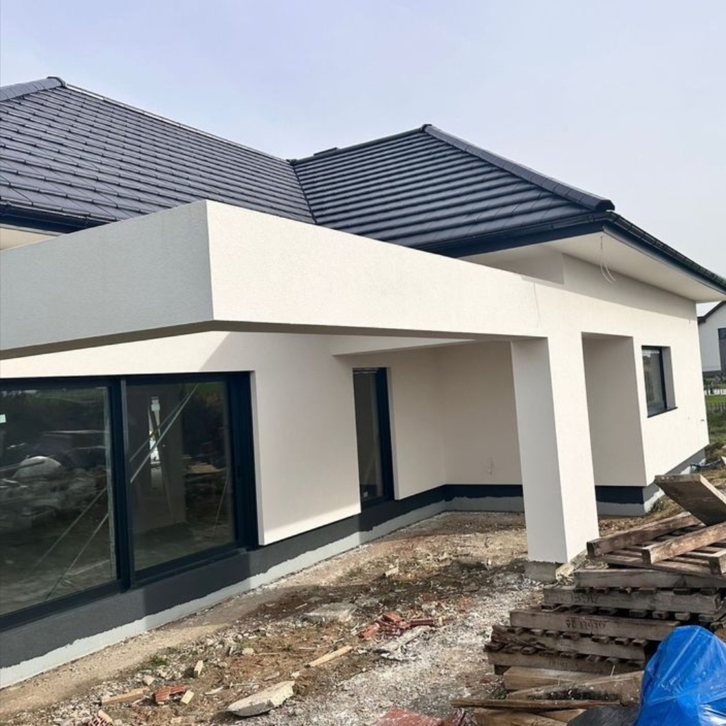 Biała elewacja domu z ciemną dachówką i dużymi oknami, widok z zewnątrz. Fragment placu budowy z drewnianymi elementami w tle. Minimalistyczny design.