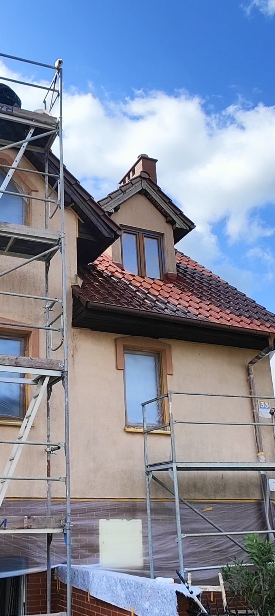 Dom jednorodzinny w trakcie renowacji elewacji. Widoczne rusztowania, zabezpieczone okna i fragment nowej, ceglanej elewacji. Niebo z białymi chmurami w tle.