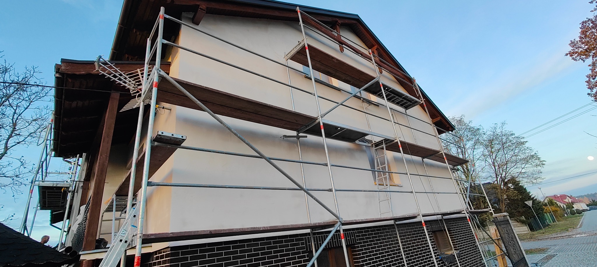 Dom jednorodzinny w trakcie malowania elewacji, widoczne rusztowania i świeżo pomalowana ściana w jasnym odcieniu. Dolna część elewacji wykończona ciemną cegłą.