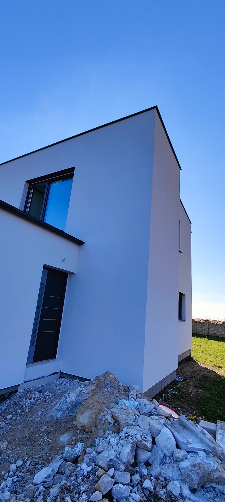 Nowoczesny, biały dom jednorodzinny z płaskim dachem, widziany z boku, z gruzem budowlanym u podstawy. Błękitne niebo w tle, podkreślające minimalistyczny design.