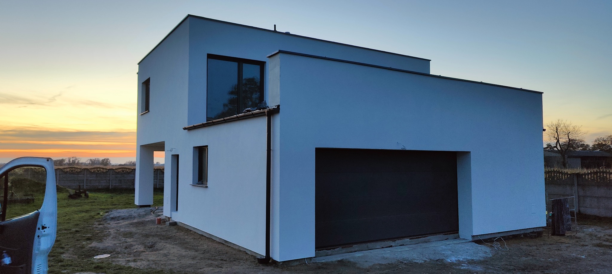 Nowoczesny, dwukondygnacyjny dom z płaskim dachem i garażem, świeżo po malowaniu elewacji na biało, uchwycony o zmierzchu z widocznym fragmentem samochodu.