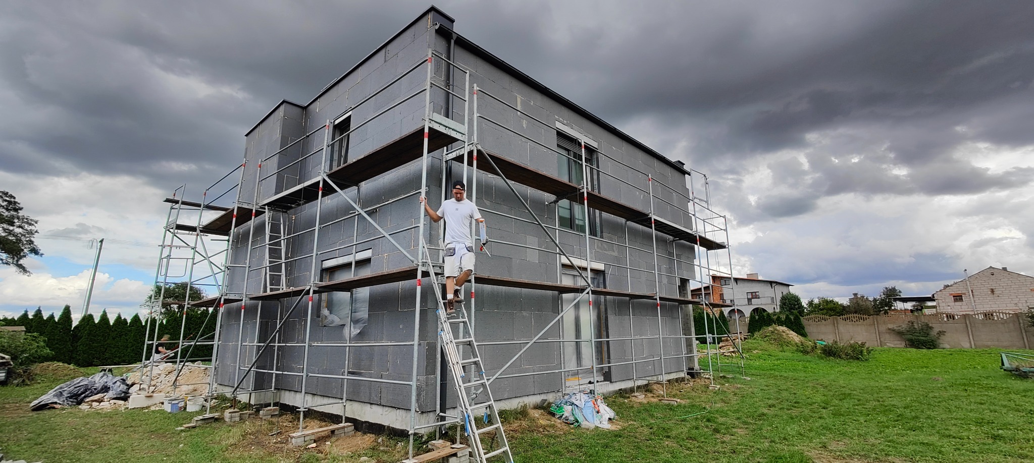 Dom w trakcie ocieplania, pokryty szarym styropianem, otoczony rusztowaniami. Pracownik na drabinie z tubą kleju. Pochmurne niebo w tle.