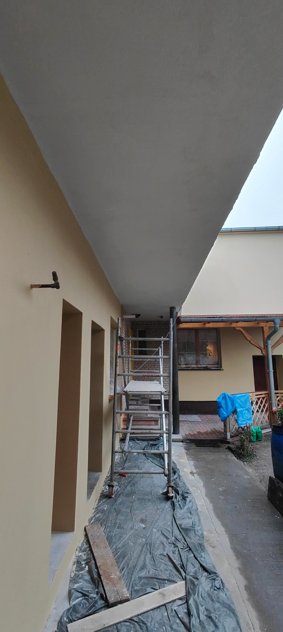 Świeżo pomalowane podbitki w beżowym odcieniu, widoczne rusztowanie i zabezpieczona folią podłoga, sugerujące prace wykończeniowe na zewnątrz budynku.