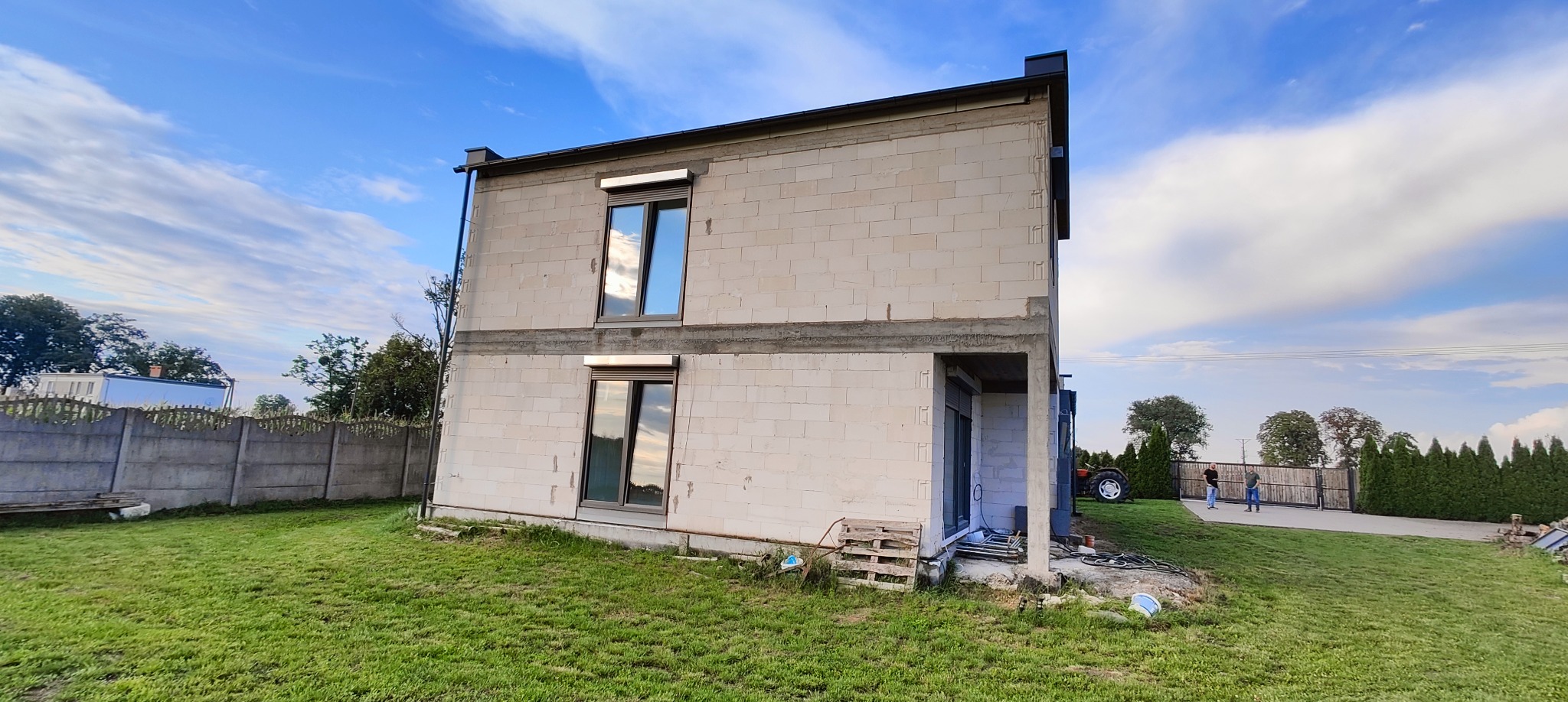 Dwukondygnacyjny dom w stanie surowym otwartym, z widocznymi oknami z roletami zewnętrznymi i niezakończoną elewacją z betonu komórkowego. Na trawniku paleta i narzędzia.