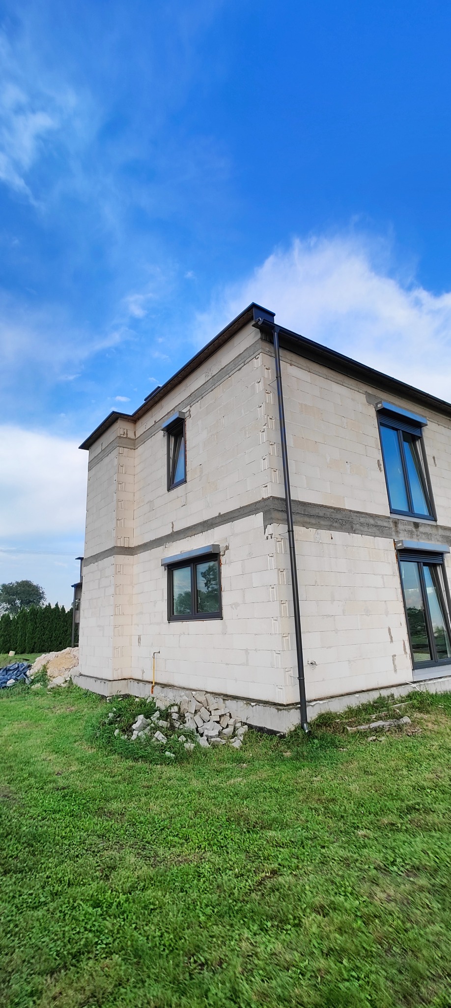 Surowy budynek z betonu komórkowego z zamontowanymi oknami i rynnami, widoczny na tle błękitnego nieba z chmurami.