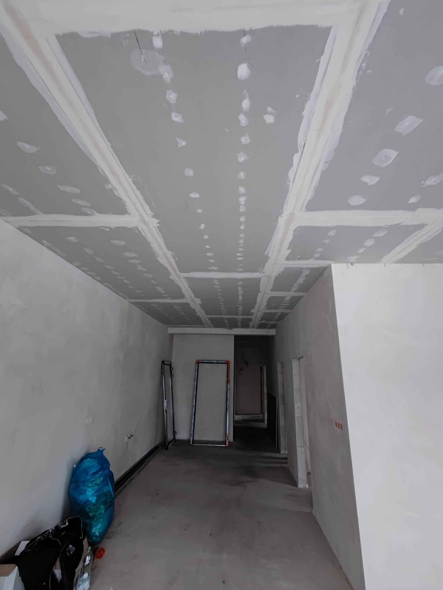 Sufit z płyt kartonowo-gipsowych, świeżo po szpachlowaniu, z widocznymi łączeniami i punktowymi śladami po szpachli. W tle widoczne ściany w trakcie prac wykończeniowych.