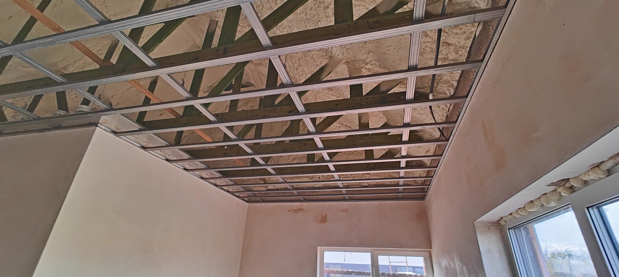 Sufit podwieszany z metalową konstrukcją i izolacją z pianki, widok z dołu w surowym wnętrzu. Ściany przygotowane do malowania, okno z pianką montażową.