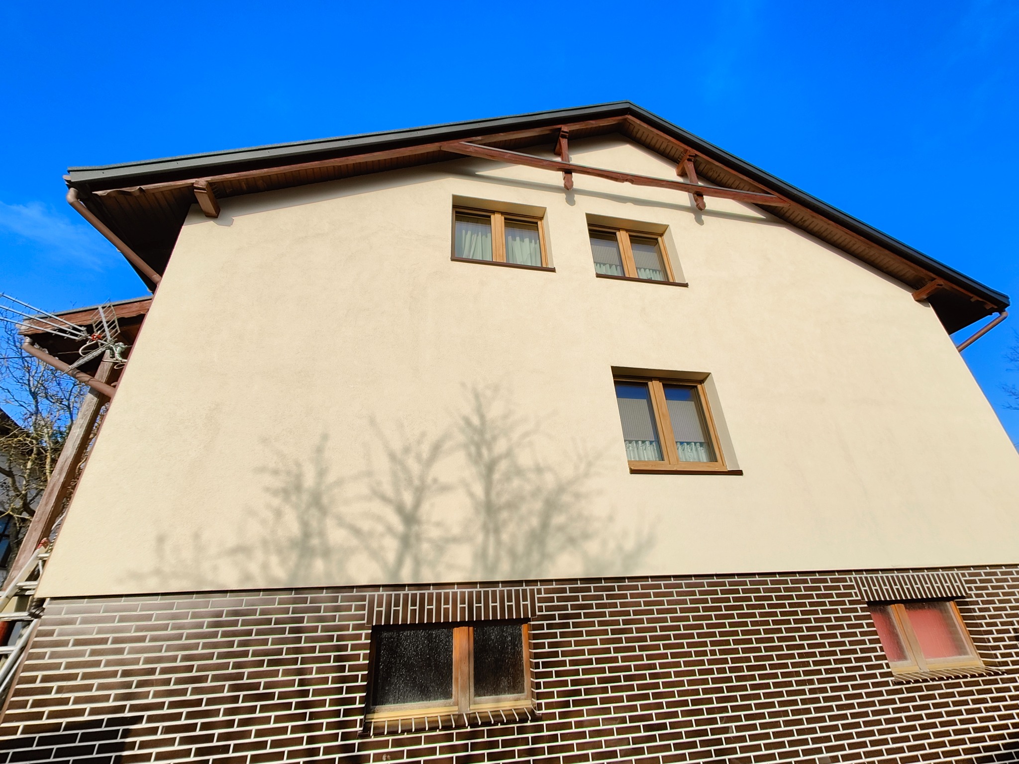 Elewacja domu z beżowym tynkiem i ceglaną podmurówką, okna z drewnianymi ramami, cień drzewa na ścianie, drabina oparta o budynek, błękitne niebo w tle.