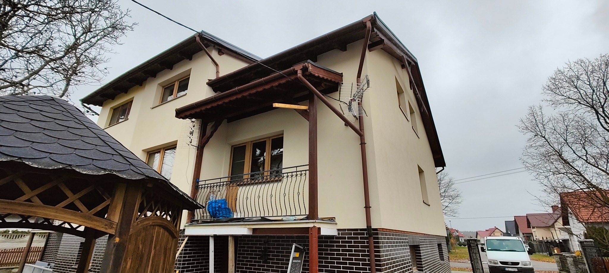 Elewacja domu po renowacji z brązowymi elementami wykończeniowymi, widoczna antena i fragment altany z boku. Ujęcie z dołu, pochmurne niebo.