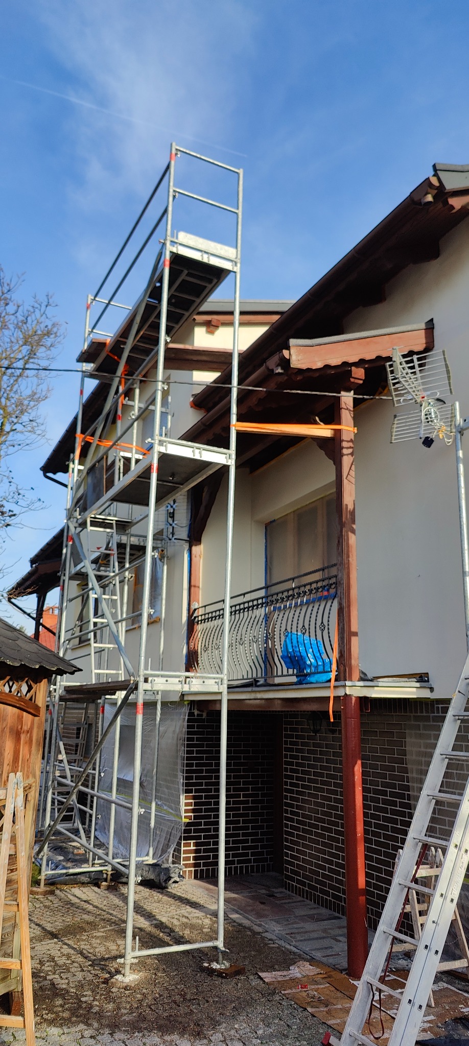 Dom jednorodzinny w trakcie prac elewacyjnych; widoczne rusztowania, zabezpieczone okna i świeżo pomalowana ściana. Ujęcie z dołu, błękitne niebo w tle.