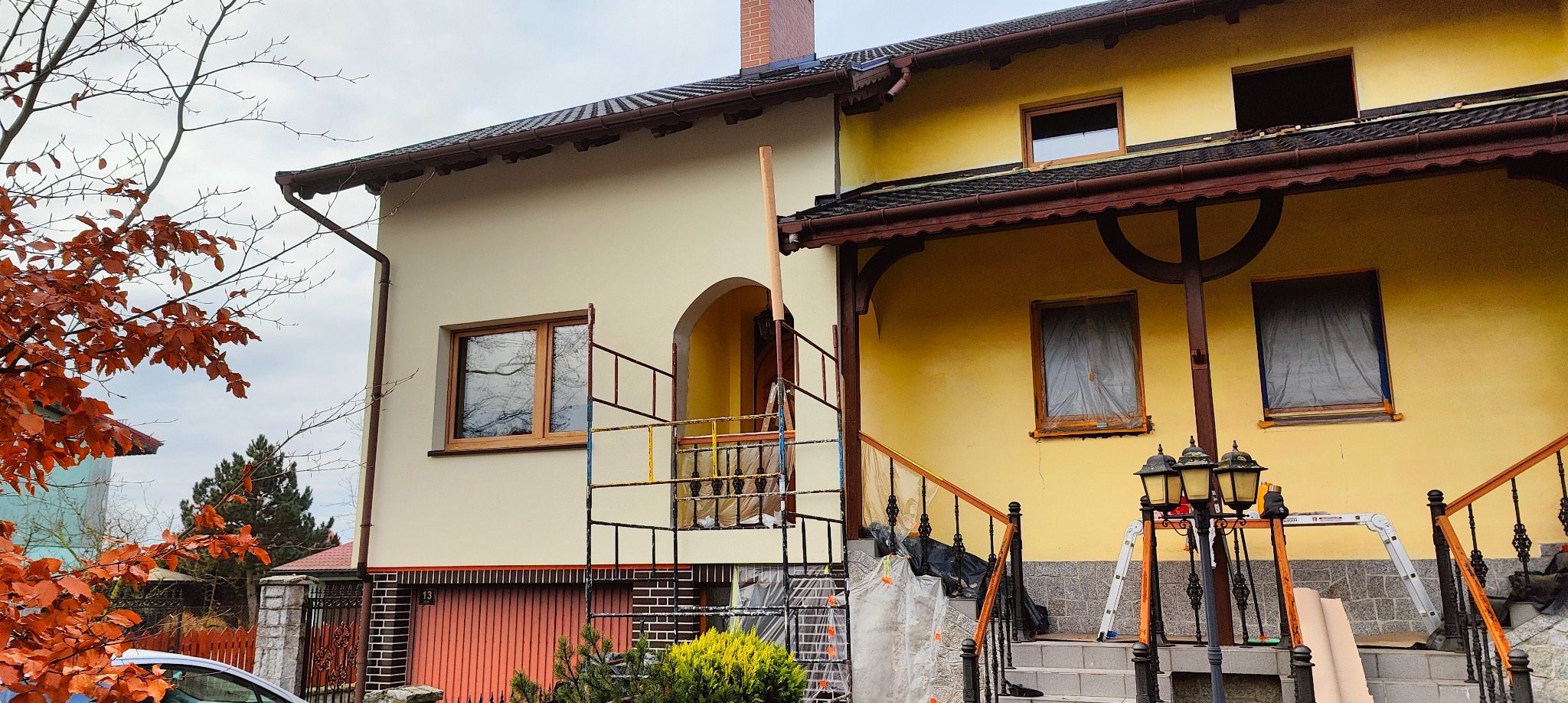 Dom dwukolorowy w trakcie renowacji elewacji: jedna część kremowa, druga żółta, rusztowania przy wejściu, okna zabezpieczone folią. Widok z zewnątrz.