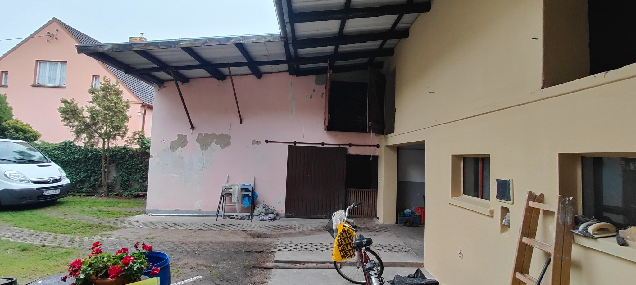 Budynek z fragmentami elewacji w trakcie malowania na beżowo, widoczne narzędzia malarskie i rower. Część budynku w kolorze różowym z odpadającym tynkiem.