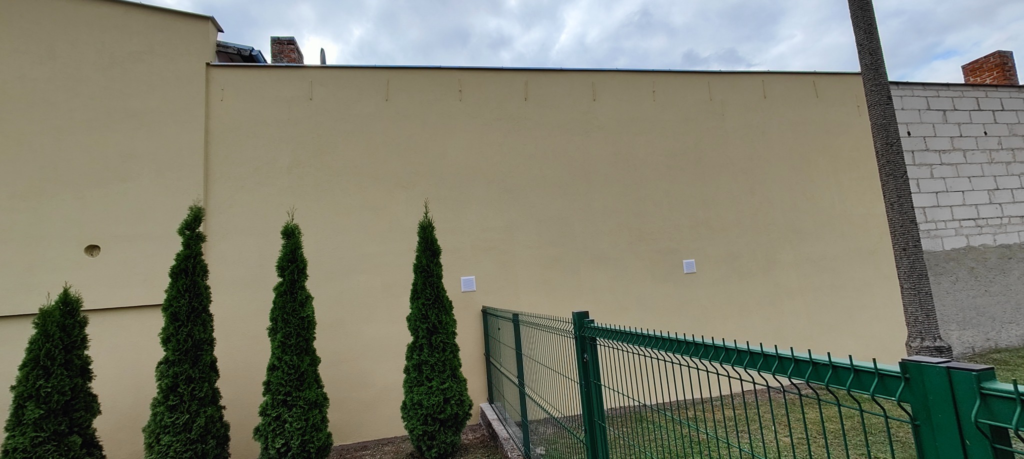 Jasna elewacja budynku z trzema tujami i zielonym ogrodzeniem. Widoczne detale: otwór w ścianie, kratki wentylacyjne. Pochmurne niebo w tle.