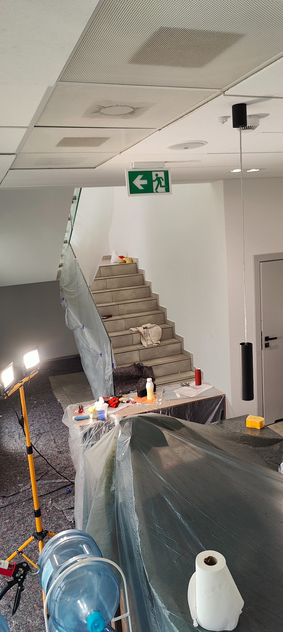 Przygotowania do malowania klatki schodowej: zabezpieczone schody folią, narzędzia na stole, oświetlenie robocze. Widoczny znak ewakuacyjny.
