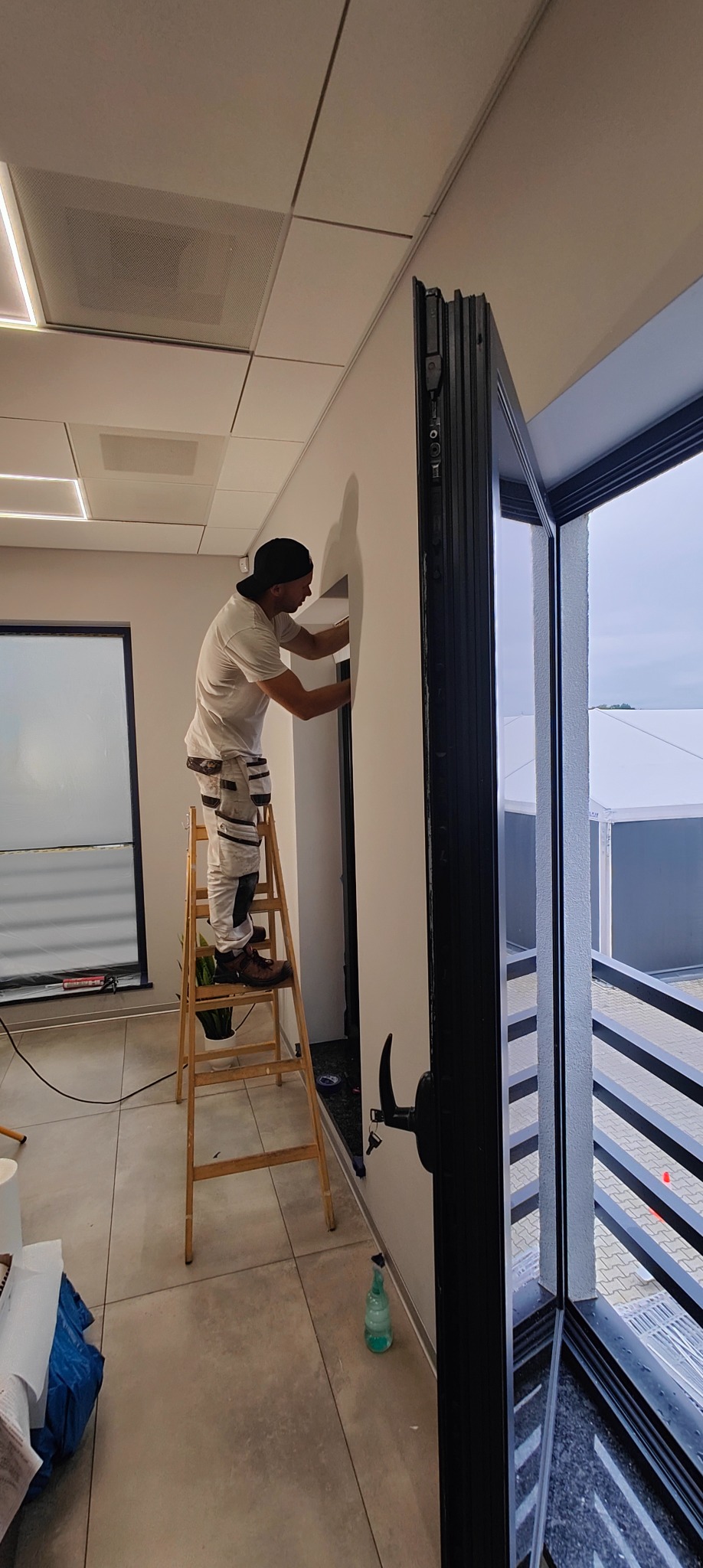 Pracownik na drabinie szpachluje ścianę w jasnym wnętrzu, widoczne okno balkonowe i sufit kasetonowy. Praca w trakcie wykonywania, detale wykończeniowe.