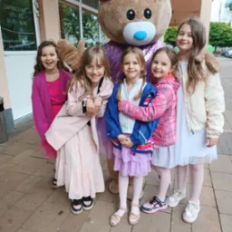 Uśmiechnięte dziewczynki pozują z animatorem przebranym za dużego misia z fioletową kokardą. Dziewczynki ubrane w różowe, białe i fioletowe sukienki i kurtki.