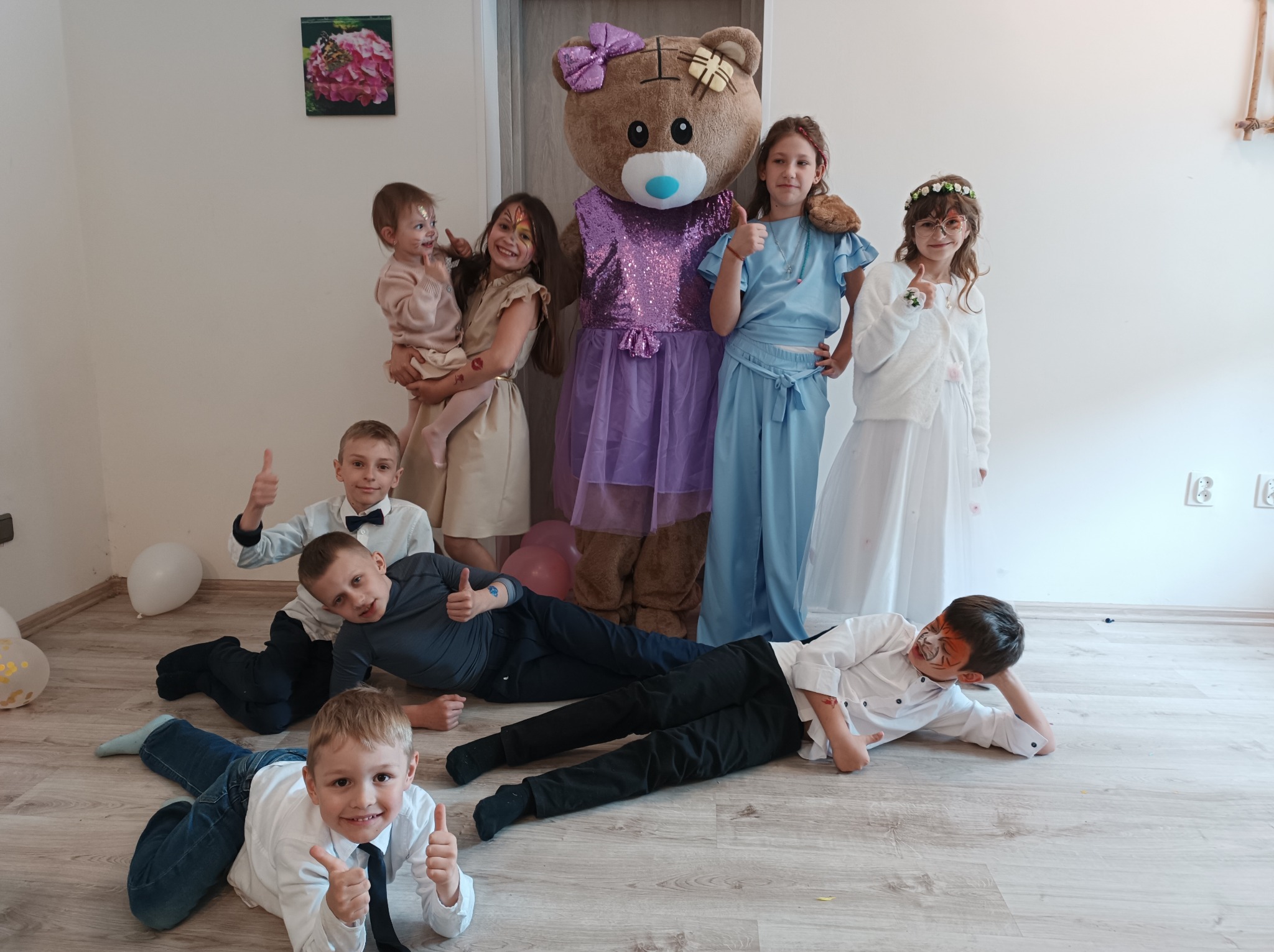 Animator w stroju misia z dziećmi z pomalowanymi twarzami, leżącymi na podłodze i pokazującymi kciuki w górę, Łódź.