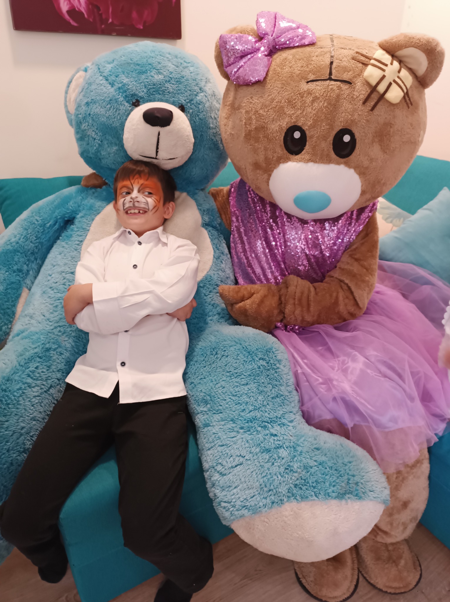 Chłopiec z pomalowaną twarzą tygrysa pozuje z pluszowymi misiami w kostiumach na niebieskiej kanapie, uśmiechając się do zdjęcia.