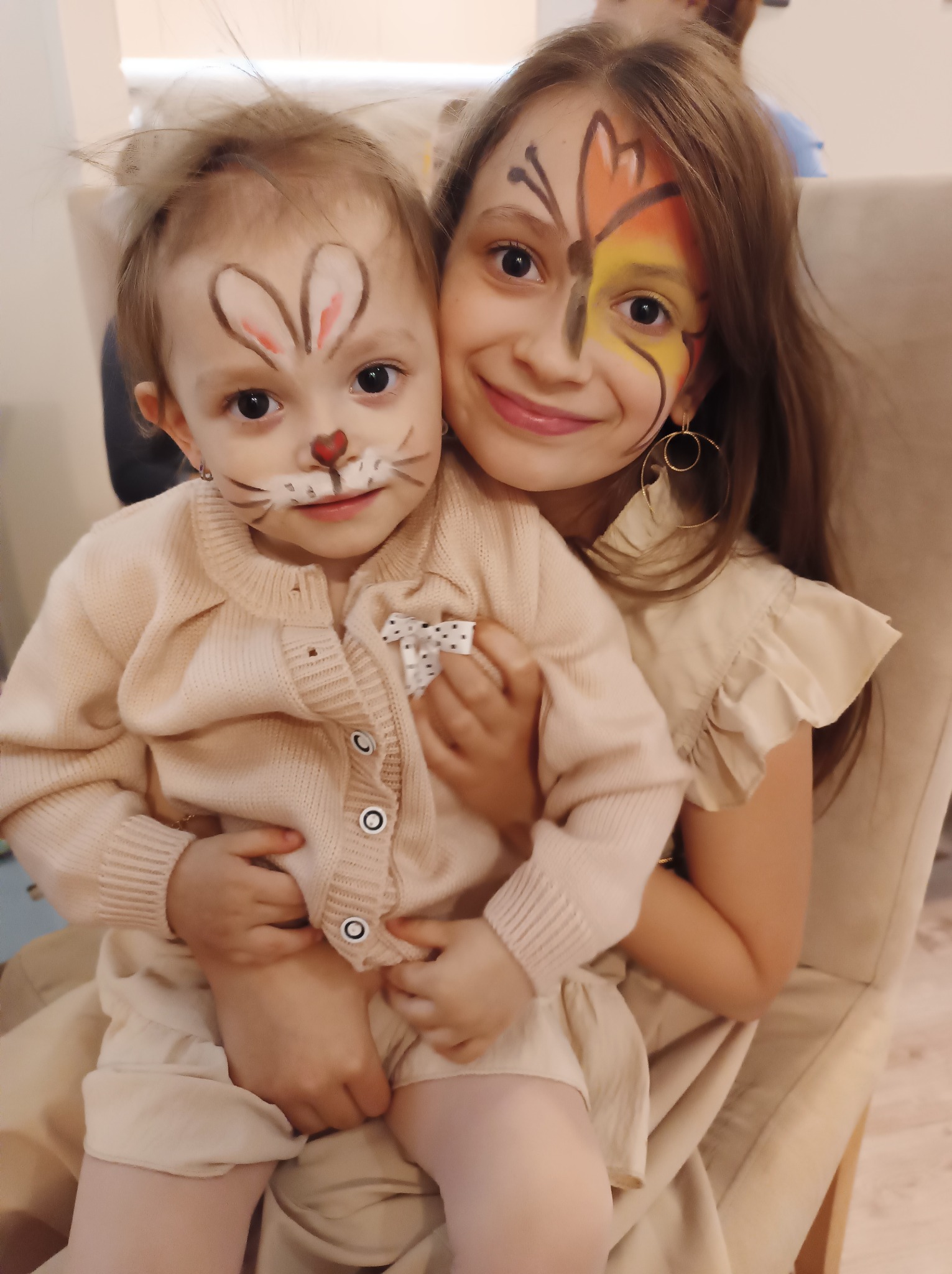 Uśmiechnięte dziewczynki z pomalowanymi twarzami: młodsza jako królik, starsza z motylem na policzku. Makijaż artystyczny w odcieniach beżu, pomarańczy i żółci.