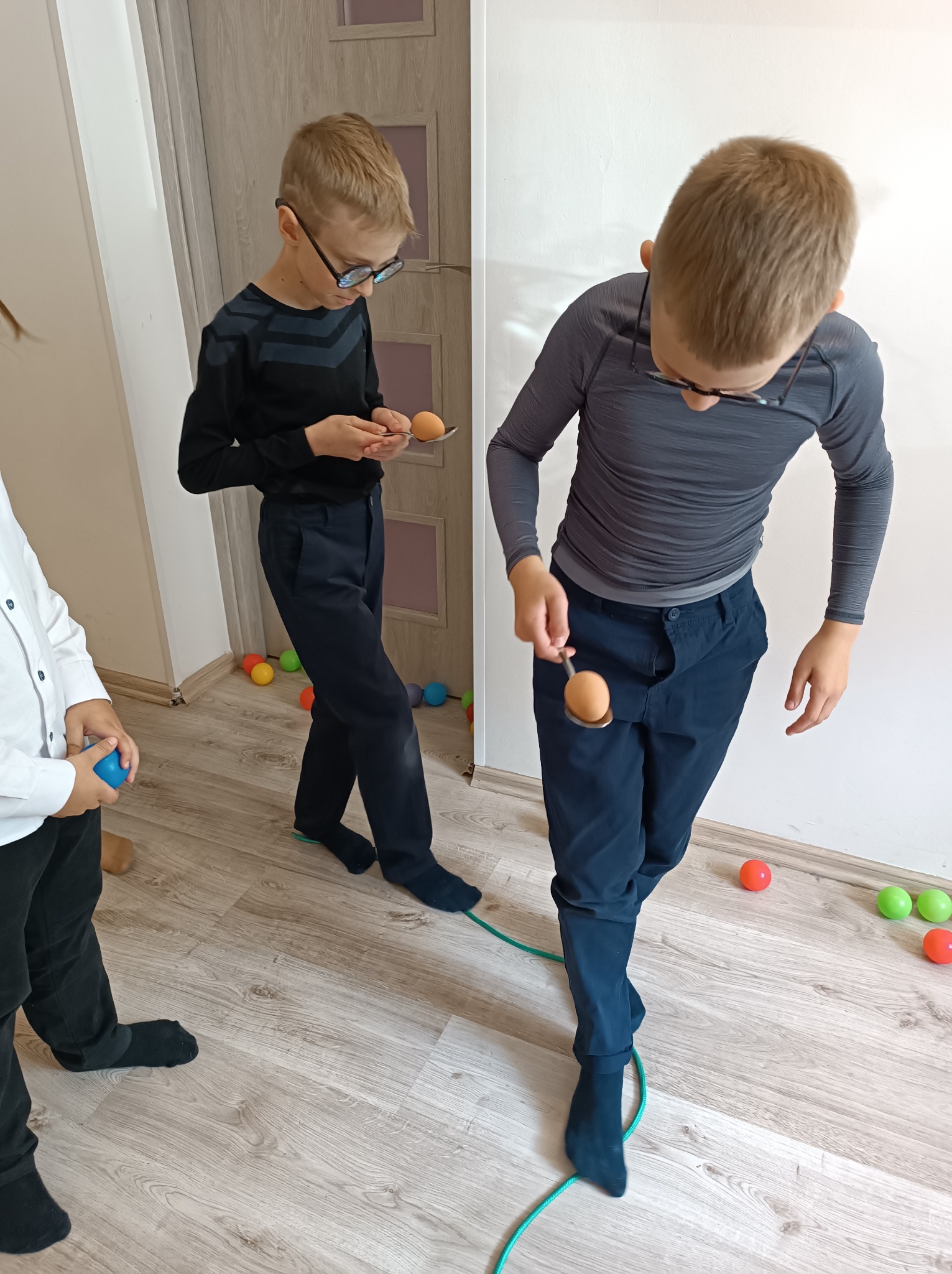 Dzieci w trakcie zabawy: przenoszenie jajka na łyżce, balansowanie, kolorowe piłki rozsypane na podłodze, elementy wyposażenia wnętrza.