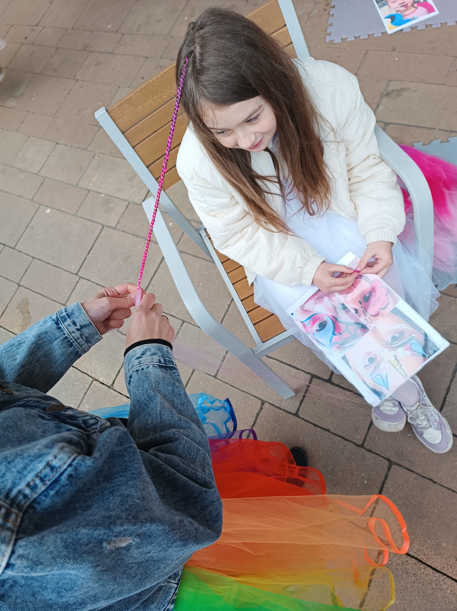 Dziewczynka siedzi na krześle, a animator zaplata jej kolorowe warkoczyki. Na kolanach ma wzornik do malowania twarzy i tęczowe worki na prezenty.